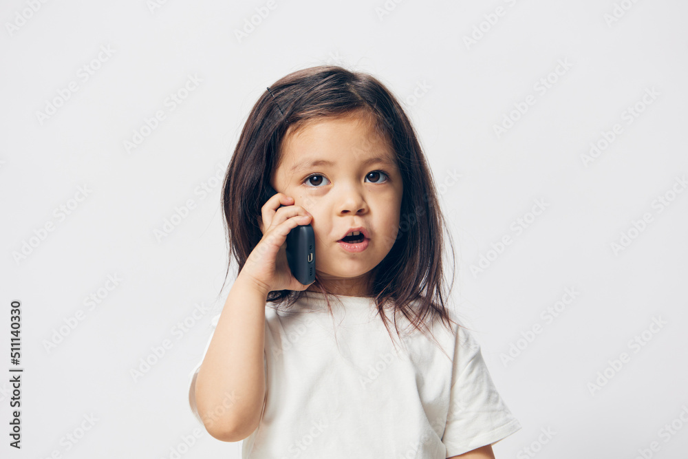a little girl of preschool age, sitting on a white background in a white T-shirt, enthusiastically talking on the phone, turning half-side to the camera. The topic of the conversation on the phone a little girl of preschool age, sitting on a white background in a white T-shirt, enthusiastically talking on the phone, turning half-side to the camera. The topic of the conversation on the phone