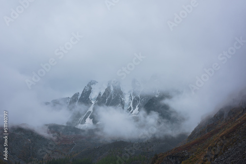 Dark atmospheric landscape with high snow mountains in dense fog in rainy weather. Large snowy mountain range in thick fog in dramatic overcast. Snow-covered rocky mountains in low clouds during rain. © Daniil