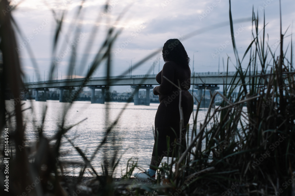 Curly African American black-haired mixed race woman walking near sea coast and bridge outside. Interracial female, summer weekend, vacation. Dusk cloud sky horizon on background. Plants foreground Curly African American black-haired mixed race woman walking near sea coast and bridge outside. Interracial female, summer weekend, vacation. Dusk cloud sky horizon on background. Plants foreground
