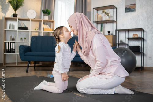 Happy Muslim family, young smiling mother and her adorable little daughter relaxing after sports sitting on the mat and touching their noses in the living room at home.