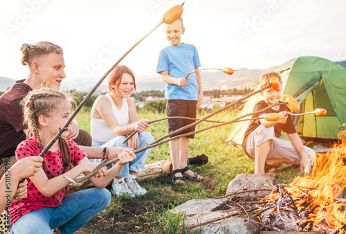 Wallpaper Mural Group of six Kids: Boys and girls cheerfully smiling and roasting sausages on sticks over a campfire flame near the green tent. Outdoor active time spending or camping in Nature concept. Torontodigital.ca