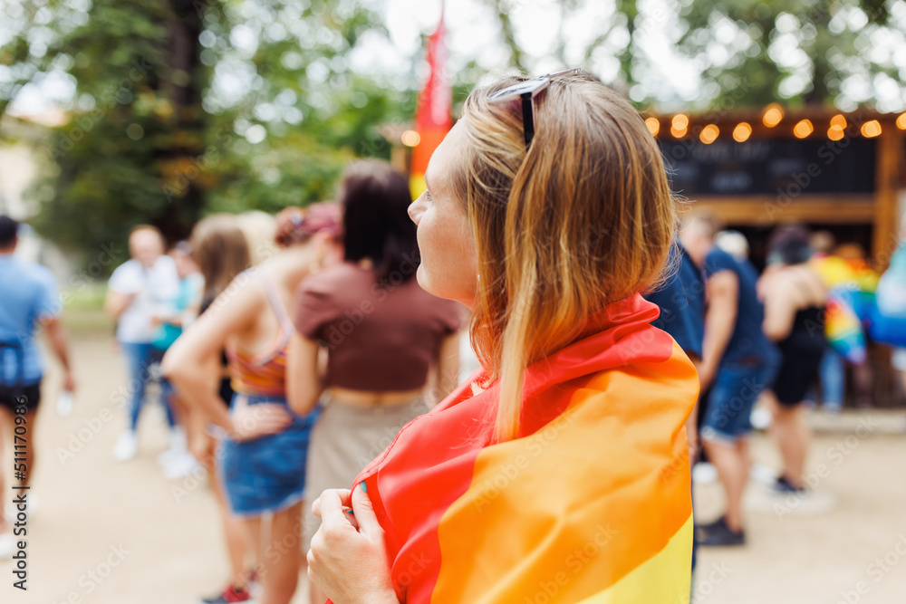 Happy young blonde hair woman covering with LGBTQI pride rainbow flag on the street in the park. LGBT Pride Month, pride event and Gay parade concept