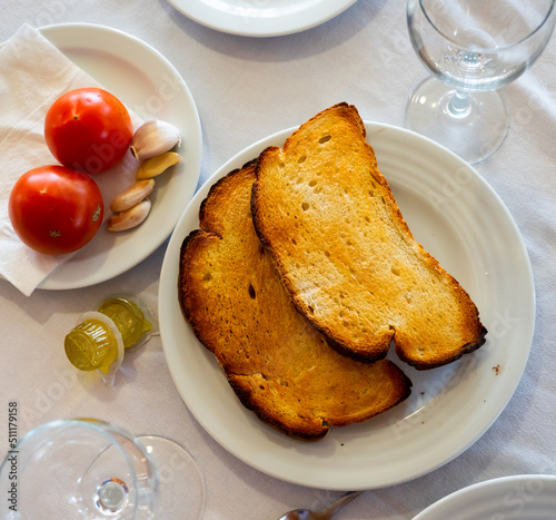 Ingredients for traditional Spanish snack Pa amb tomaquet served on table. Crispy toasted bread, fresh tomatoes and garlic on plates and olive oil in individual packages photo
