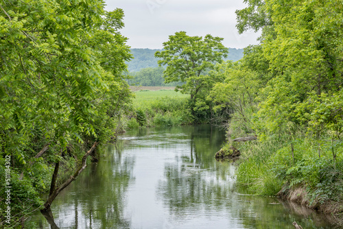 A river running through farm country with trees on the banks and a wooded hill in the background in the summer.