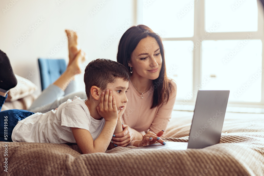 Attractive Caucasian mother and small teen son lying on bed and looking at laptop screen.