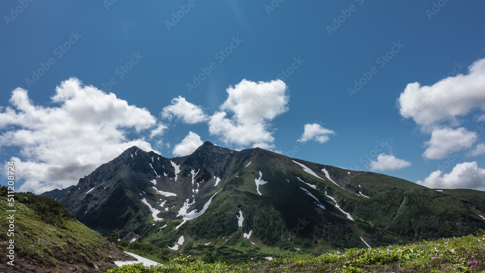 A picturesque mountain range against a background of blue sky and clouds. Melted snow on the slopes. Green vegetation on an alpine meadow. Kamchatka. Vachkazhets. 
