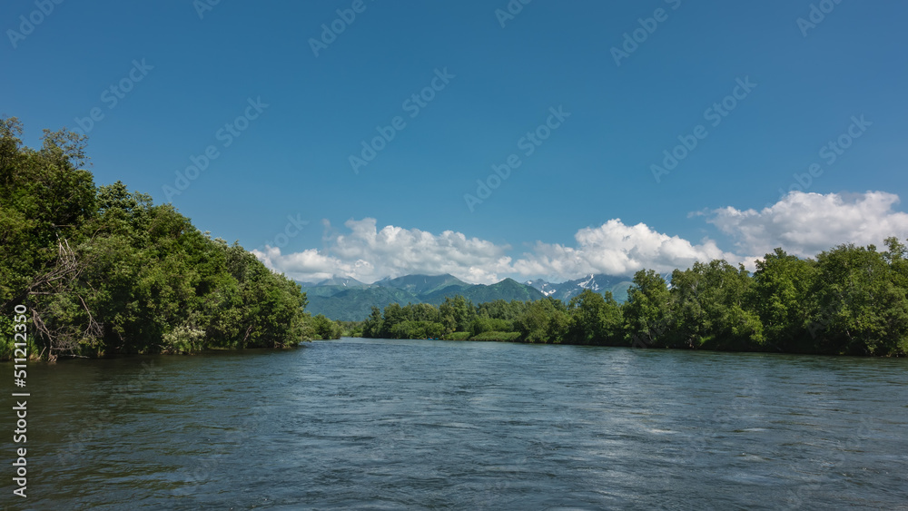 A calm blue river under an azure sky. There are thickets of green trees on the banks. A mountain range in the clouds. Kamchatka. River Bystraya