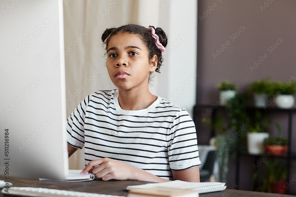 Serious African girl concentrating on online meeting while sitting at table in front of computer monitor during her remote studying Serious African girl concentrating on online meeting while sitting at table in front of computer monitor during her remote studying