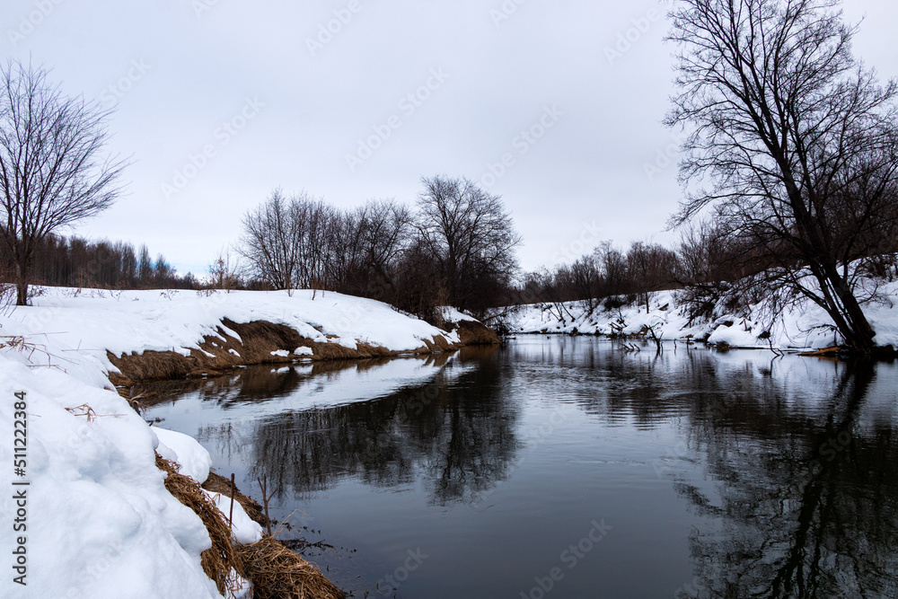 forest river with fast flow and small waves flows between snowy shores with trees and bushes late winter and early spring forest river with fast flow and small waves flows between snowy shores with trees and bushes late winter and early spring