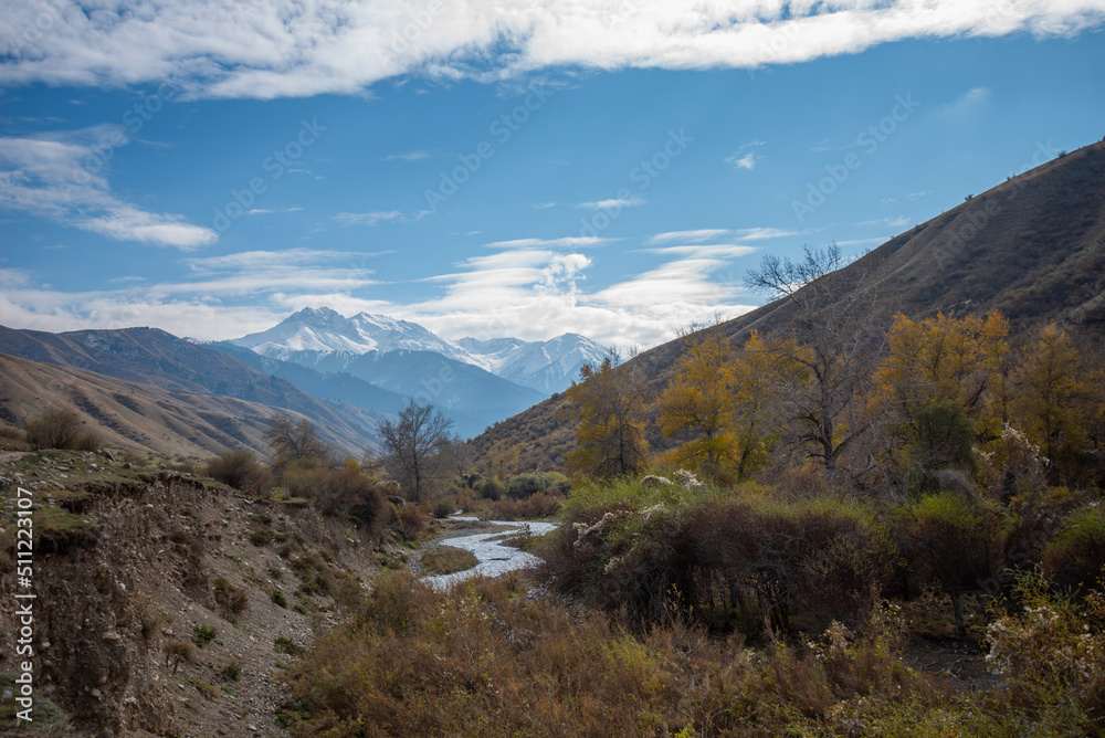 Mountain valley with a small river and a country road. A mountain range with snow-capped peaks in a blue haze in the distance. Bright autumn sky with beautiful clouds and yellow trees.