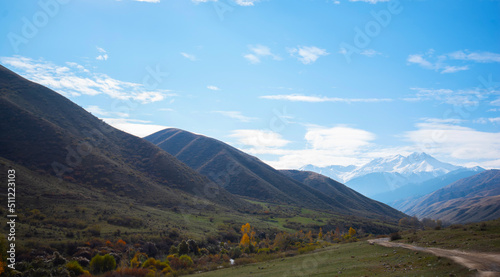 Selective focus. Mountain valley with a small river and a country road. A mountain range with snow-capped peaks in a blue haze in the distance. Autumn sky with beautiful clouds and yellow trees.