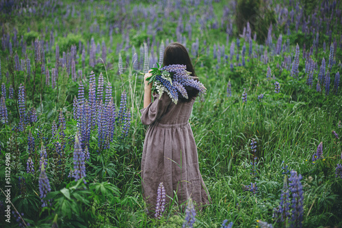 Cottagecore aesthetics. Stylish woman in rustic dress gathering lupine bouquet in meadow. Young female in linen dress with wildflowers in atmospheric summer countryside, rural slow life