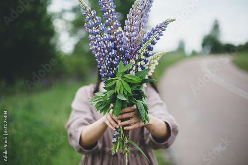Lupine bouquet in woman hands close up in summer countryside. Cottagecore aesthetics. Young female in linen dress holding wildflowers on background of rural road and meadow, slow life