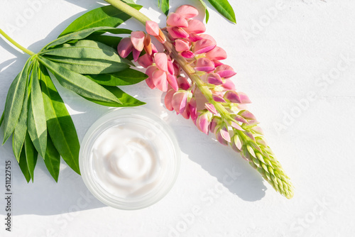 A white cosmetic jar with face natural cream on a white background next to pink lupine and green leaves. Natural organic skin care cosmetics