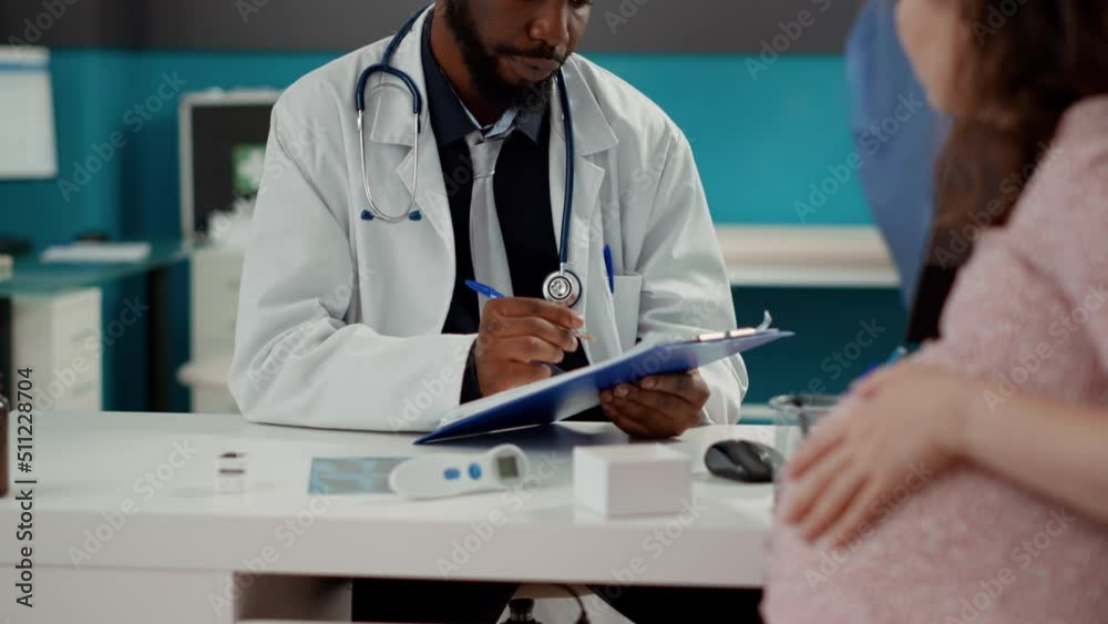 African american medic taking notes at consultation with pregnant person, writing checkup report on documents. Woman with pregnancy belly asking for maternity advice from male obstetrician. African american medic taking notes at consultation with pregnant person, writing checkup report on documents. Woman with pregnancy belly asking for maternity advice from male obstetrician.