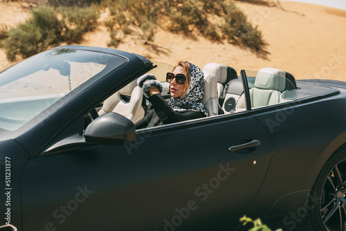 Glamorous blonde in a silk scarf and sunglasses in the driver's seat of a convertible convertible on a hot desert background. Fashion, style, glamour © Sviatlana