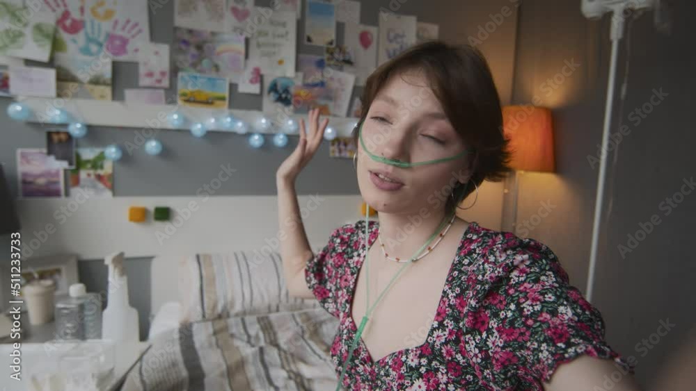 Handheld POV of short-haired Caucasian girl with nasal cannula sitting on bed in decorated hospital ward, smiling, talking on camera, showing pill jars, filming vlog Handheld POV of short-haired Caucasian girl with nasal cannula sitting on bed in decorated hospital ward, smiling, talking on camera, showing pill jars, filming vlog