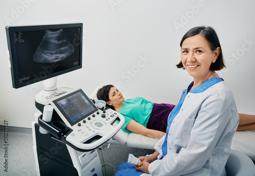 Portrait of sonographer near the ultrasound machine at medical clinic during female patient's body ultrasound scan. Sonographer occupation photo