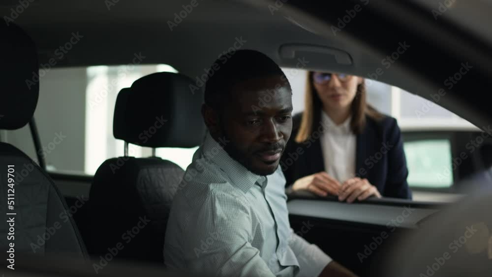 Young businessman is sitting inside car and talking to salesperson in dealership office spbas. 4k Close view of bearded African man examines new car and looks with smile, consults with female manager Young businessman is sitting inside car and talking to salesperson in dealership office spbas. 4k Close view of bearded African man examines new car and looks with smile, consults with female manager