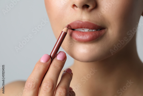 Young woman applying beautiful nude lip pencil on light grey background, closeup
