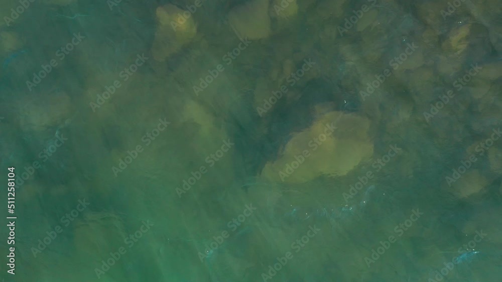 A dynamic close up shot of a blue-green colored river while seeing glares of the sun in every wave and the riverbed beneath it. The rocks are quite visible because of the clearness of the water.