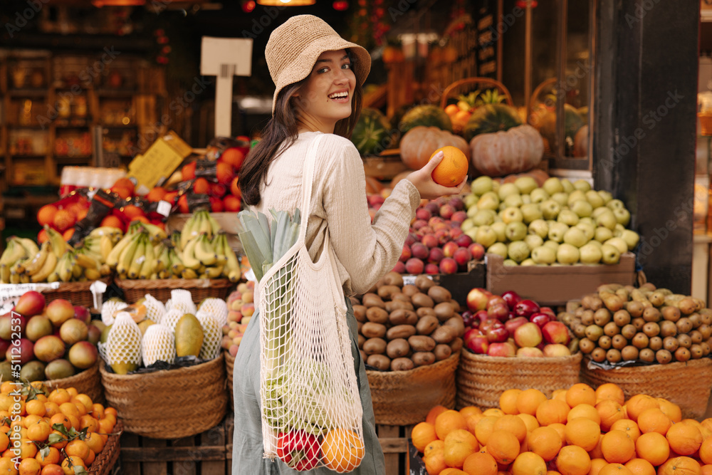 Beautiful young caucasian woman smiling turning to camera standing near fruit stand. Brunette wears casual clothes and shopping bag with groceries. Healthy food concepts