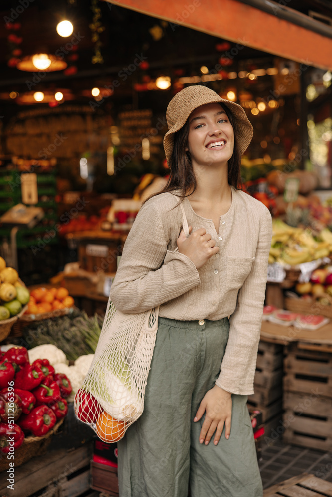 Pleasant young caucasian girl smiling looking at camera spending time at vegetable market. Brunette wears blouse, pants, hat and string bag. Buyer concept