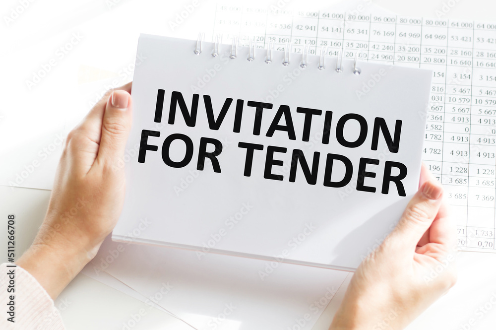 INVITATION FOR TENDER, text on white notepad in the hands of a businessman on a table next to financial reports INVITATION FOR TENDER, text on white notepad in the hands of a businessman on a table next to financial reports