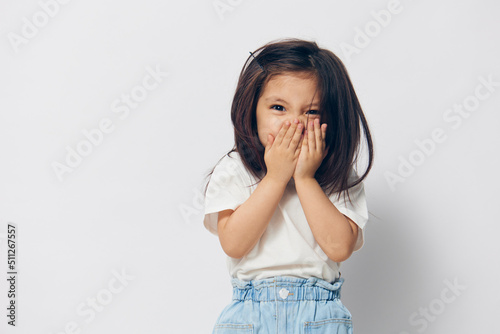 Canvas Print a little cute girl of preschool age in a white T-shirt, standing on a white background, closing her eyes with her hands and puffing out her cheeks