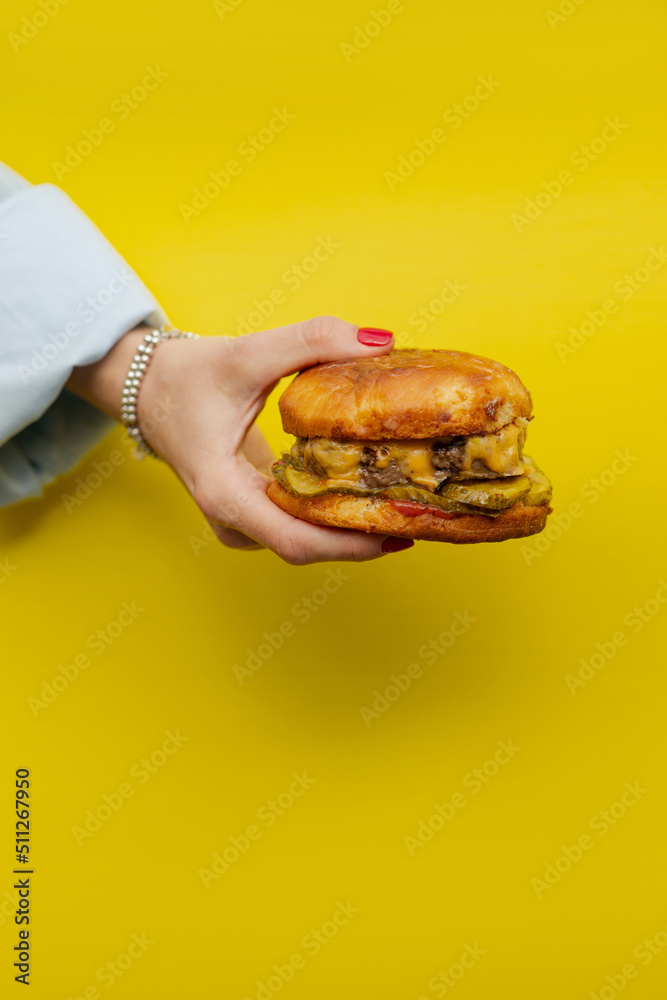 Classic cheeseburger in a woman’s hand on a bright yellow background. Juicy appetizing cheeseburger in hand and girls. Girl holding a burger close up