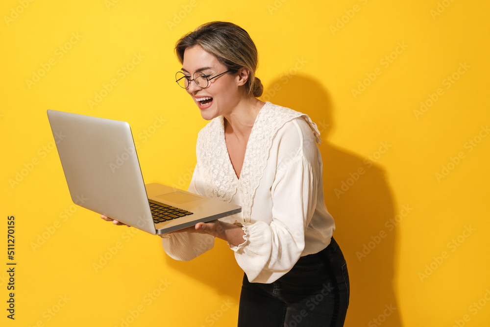 Cheerful woman wearing eyeglasses is using laptop computer on yellow background Cheerful woman wearing eyeglasses is using laptop computer on yellow background