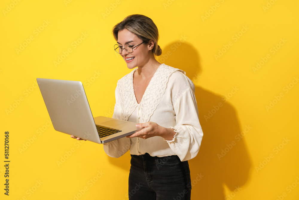 Cheerful woman wearing eyeglasses is using laptop computer on yellow background Cheerful woman wearing eyeglasses is using laptop computer on yellow background