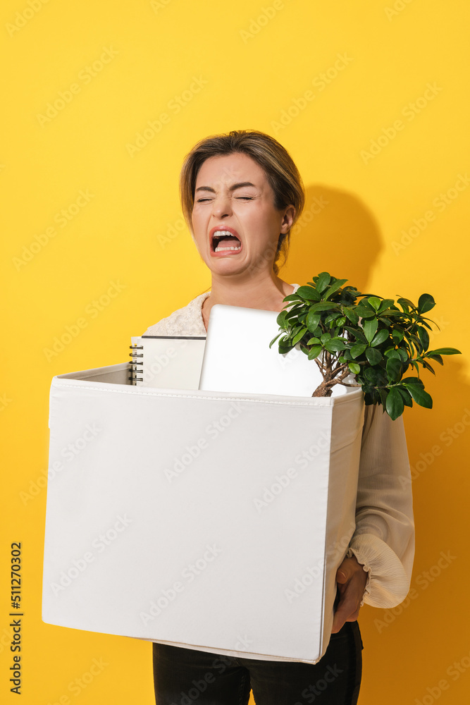Upset woman holding box with personal items after job resignation against yellow background Upset woman holding box with personal items after job resignation against yellow background