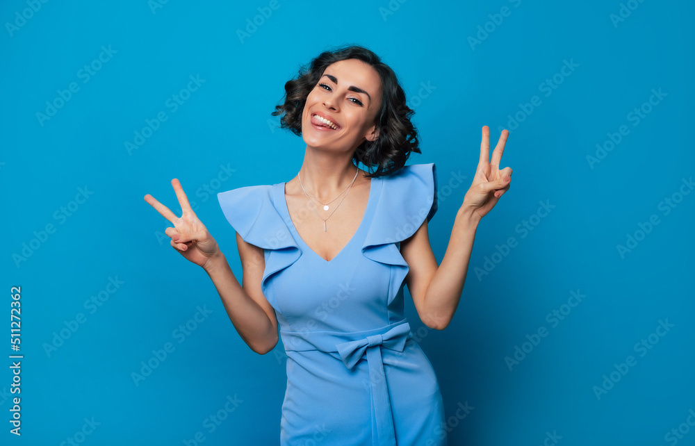 Close up portrait of an excited happy gorgeous girl in a long blue dress, who is looking in the camera while she posing with a bright toothy smile and shows victory sign on the camera Close up portrait of an excited happy gorgeous girl in a long blue dress, who is looking in the camera while she posing with a bright toothy smile and shows victory sign on the camera