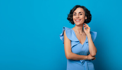 Close-up photo of a charming healthy woman in a blue dress, who is looking in the camera and smiling, while standing and posing isolated on blue background