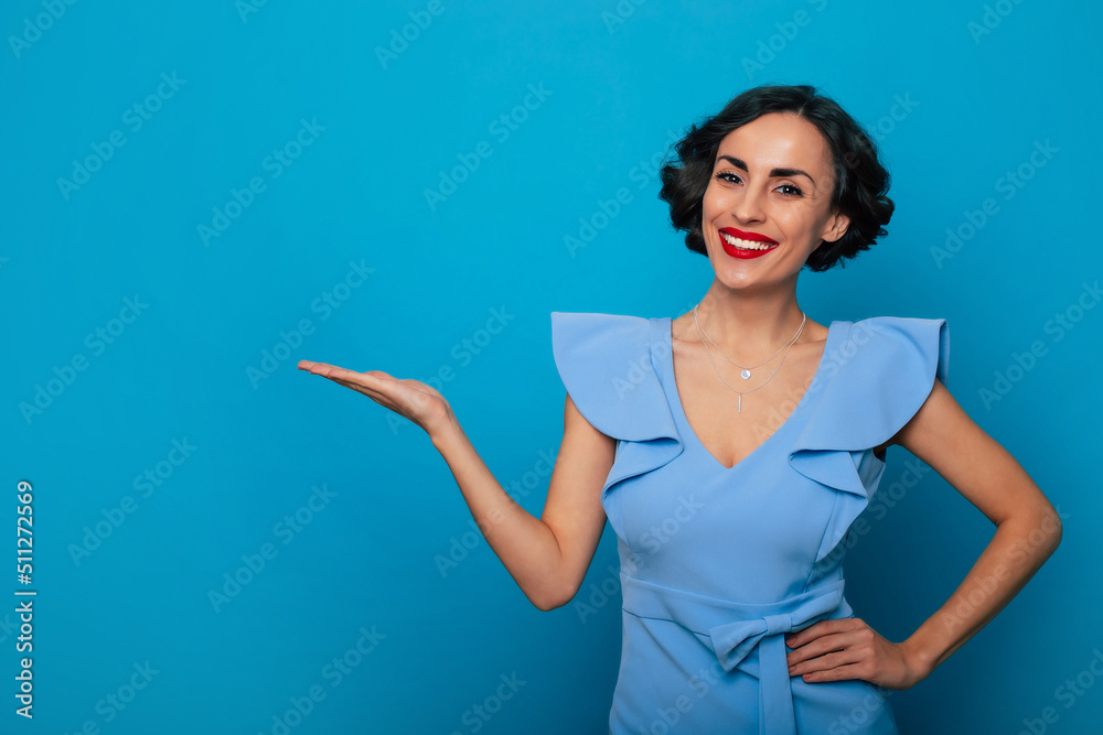 Happy attractive young brunette woman with wavy hairstyle and bright toothy smile while she shows her empty palm in dress on blue background Happy attractive young brunette woman with wavy hairstyle and bright toothy smile while she shows her empty palm in dress on blue background