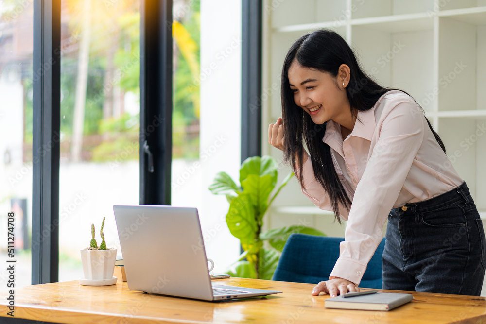 A beautiful Asian woman celebrates while using a laptop at the office and congratulates her on her business success.