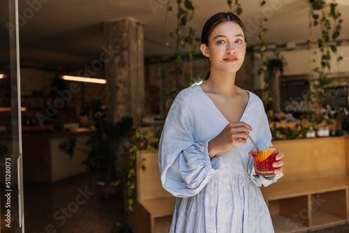 Beautiful young caucasian girl looks away, holds glass with cooling lemonade in her hands. Brunette wears blue sundress relaxing in cafe. City life concept