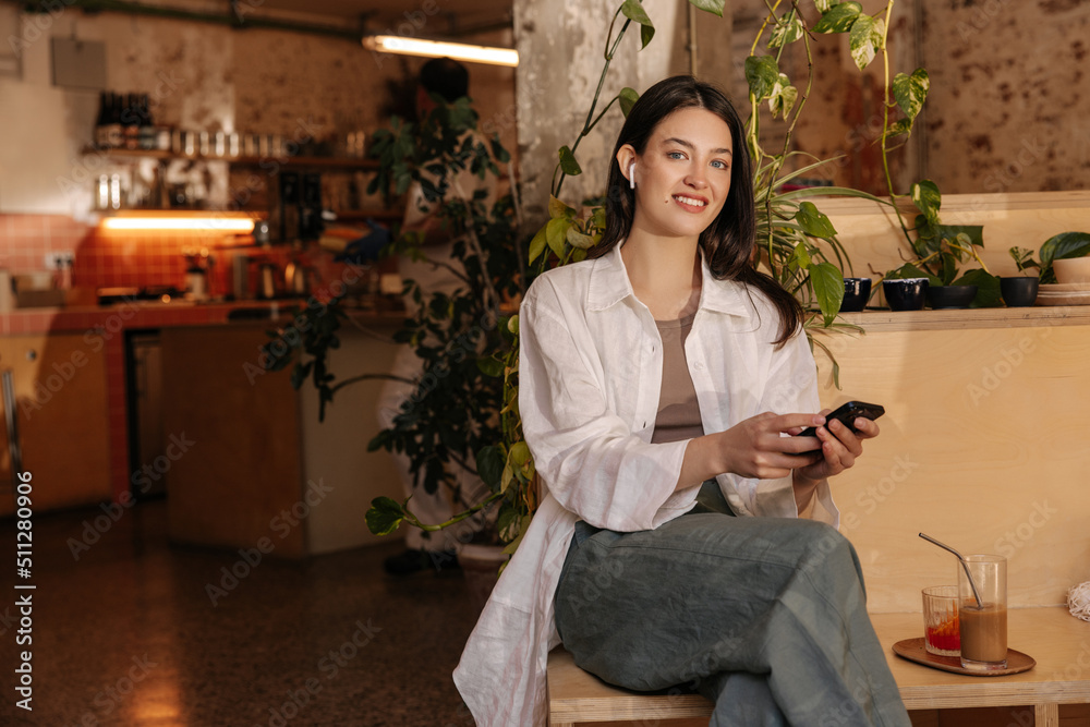 Beautiful young caucasian girl looks at camera, holds phone in hands sitting in cafe. Brunette wears casual clothes spring. Concept of use, lifestyle