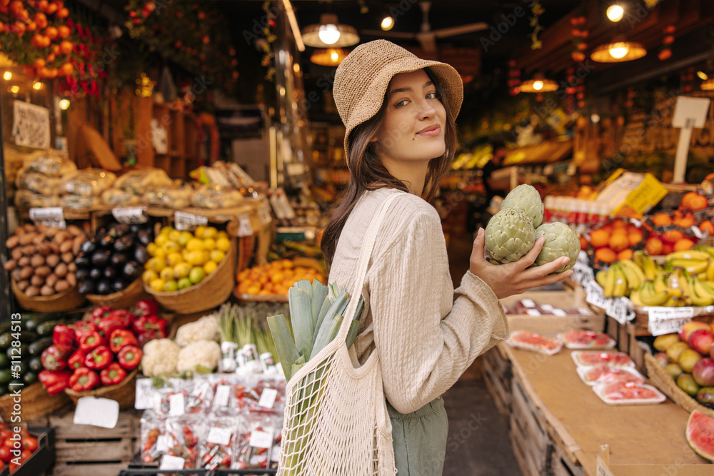 Pretty young caucasian woman looking at camera, holding fresh artichoke standing in market. Brunette wears casual clothes, hat and shopping bag. Delicious vegetables concept