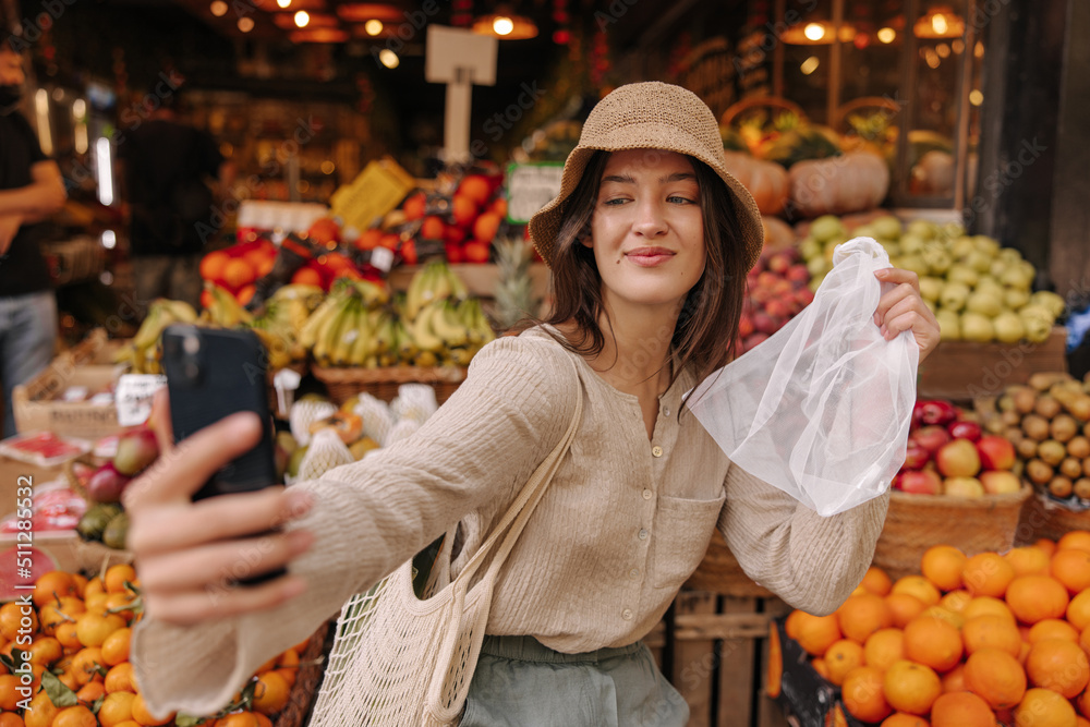 Attractive young caucasian woman is photographed on smartphone shopping for groceries. Brunette wears hat, blouse and string bag. Concept of use