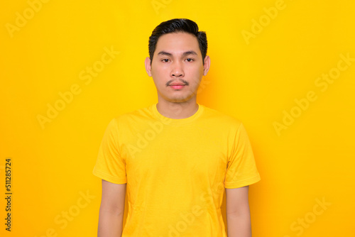 Handsome young Asian man in casual t-shirt looking at camera with confident expression isolated on yellow background. People lifestyle concept