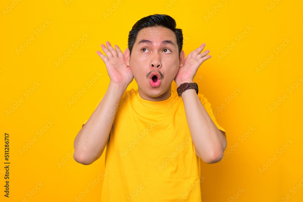 Amazed young Asian man in casual t-shirt holding hands near his ears isolated on yellow background. People lifestyle concept Amazed young Asian man in casual t-shirt holding hands near his ears isolated on yellow background. People lifestyle concept