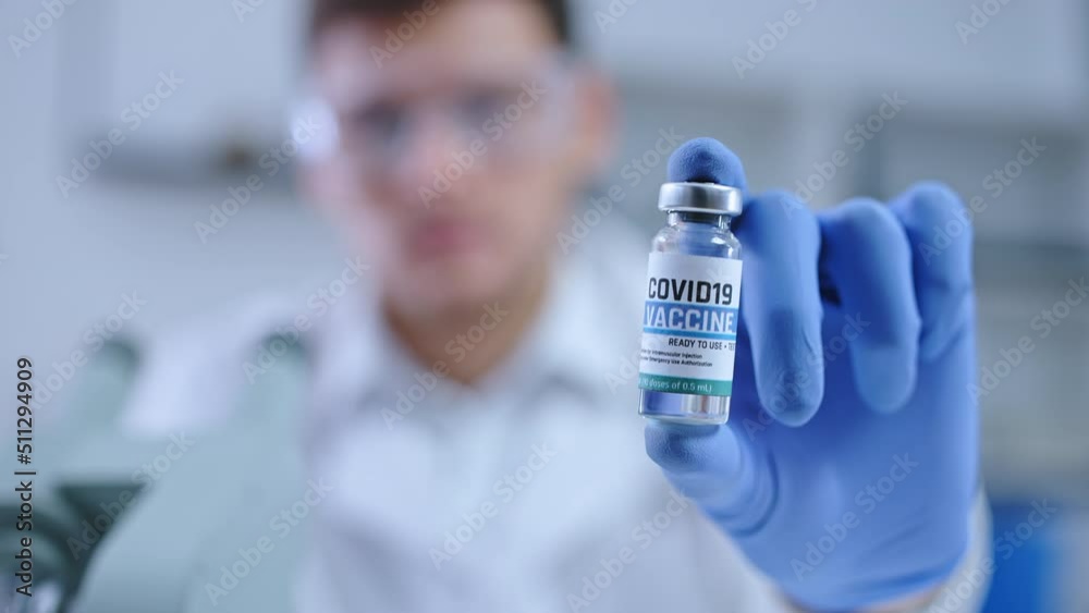 Male lab scientist in protective eyeglasses showing COVID-19 vaccine on camera Male lab scientist in protective eyeglasses showing COVID-19 vaccine on camera
