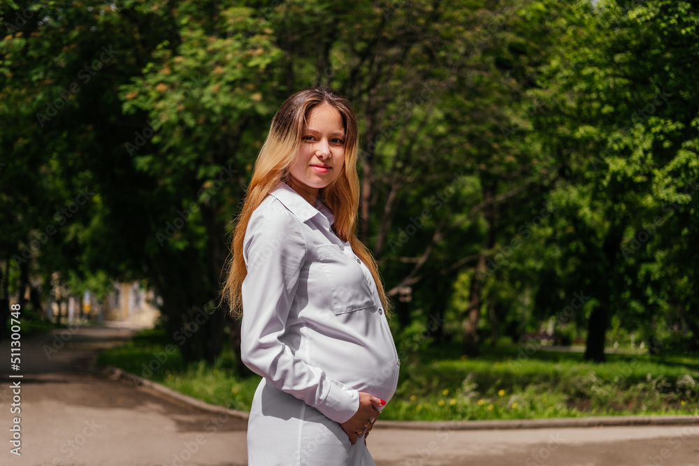 Pregnant young woman smiling and holding hand on belly looking at camera outdoors Pregnant young woman smiling and holding hand on belly looking at camera outdoors