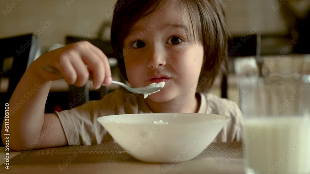 a cute Asian-looking kid is eating semolina porridge, the child is soiled with food. The boy has breakfast sitting at home in the kitchen at the table a cute Asian-looking kid is eating semolina porridge, the child is soiled with food. The boy has breakfast sitting at home in the kitchen at the table