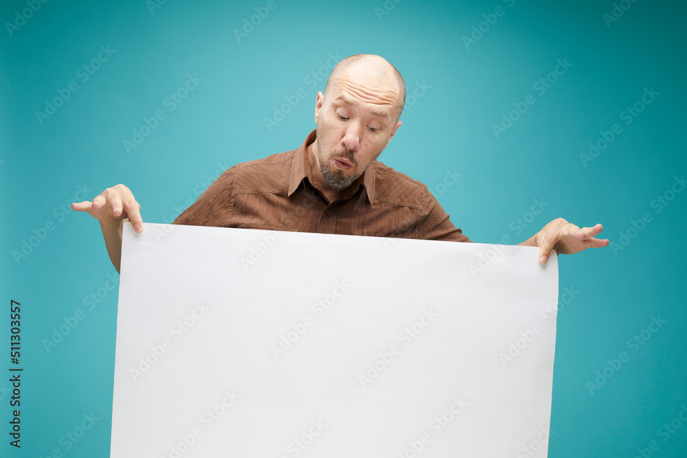 A Bald man with emotion face holding white board in his hands to fill your text. Isolated on blue background. A Bald man with emotion face holding white board in his hands to fill your text. Isolated on blue background.