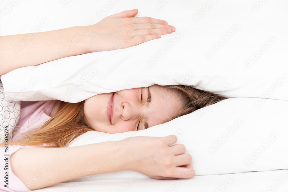 Teen girl shutting her eyes covering ears with pillows in a bed. Sleepless unhappy teenager lying in bed. Stress, relationship problems, insomnia, depression, mental health, transitional age. Teen girl shutting her eyes covering ears with pillows in a bed. Sleepless unhappy teenager lying in bed. Stress, relationship problems, insomnia, depression, mental health, transitional age.