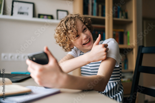 Laughing cute little girl holding and using mobile phone showing thumb up, browsing social network apps, surfing internet and scrolling news feed, reading electronic e-book at table with laptop.