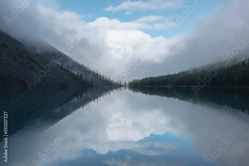 Tranquil meditative misty scenery of glacial lake with reflection of pointy fir tops and clouds at early morning. Graphic EQ of spruce silhouettes on calm alpine lake horizon. Mountain lake in fog.